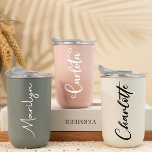 Puede incluir: Tres vasos aislados en gris, rosa y blanco, cada uno con tapa plateada. Los vasos están personalizados con los nombres "Marilyn", "Carlota" y "Charlotte" en fuente cursiva. Los vasos se exhiben sobre una superficie de madera.