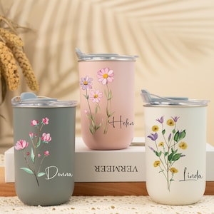 Op de afbeelding: Drie geïsoleerde bekers in grijs, roze en wit, elk versierd met bloemmotieven en gepersonaliseerde namen. De bekers hebben zilveren deksels en worden gepresenteerd op een houten oppervlak. De namen zijn Donna, Helen en Linda.