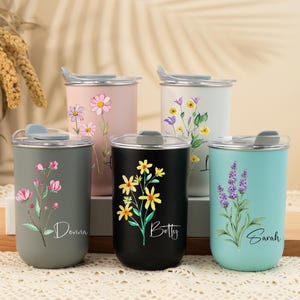 Könnte beinhalten: Fünf Edelstahl-Reisebecher in verschiedenen Farben, jeweils mit floralem Design und Namen. Die Becher sind grau, rosa, weiß, schwarz und türkis. Jeder Becher hat einen transparenten Deckel mit einem grauen Schieber.