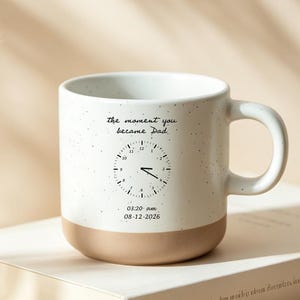 Op de afbeelding: Een witte keramische mok met een bruine basis en oor, met een klokontwerp en de tekst "the moment you became Dad". De mok staat op een wit boek op een houten plank, met een kanten kleedje en gedroogde planten op de achtergrond.