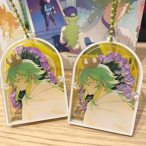 May include: Two clear acrylic charms featuring a character with green hair and a white cloak, adorned with a gold crown. They hang from a green beaded chain. A colourful illustration is visible in the background.