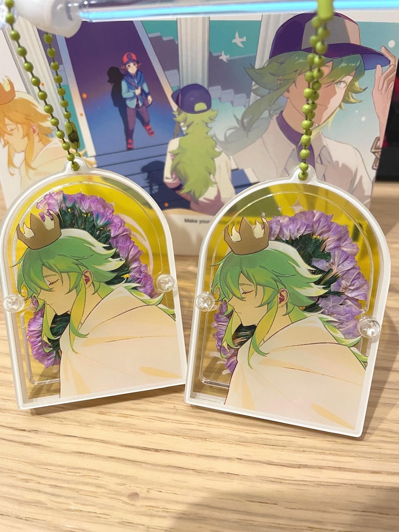 May include: Two clear acrylic charms featuring a character with green hair and a white cloak, adorned with a gold crown. They hang from a green beaded chain. A colourful illustration is visible in the background.