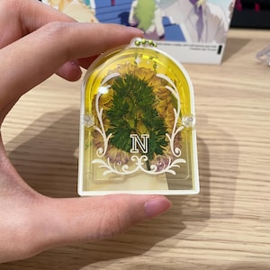 May include: A yellow and white rectangular keyring with a floral design and the letter "N". The keyring has a clear centre with dried flowers and decorative flourishes. The background shows an anime-style illustration.