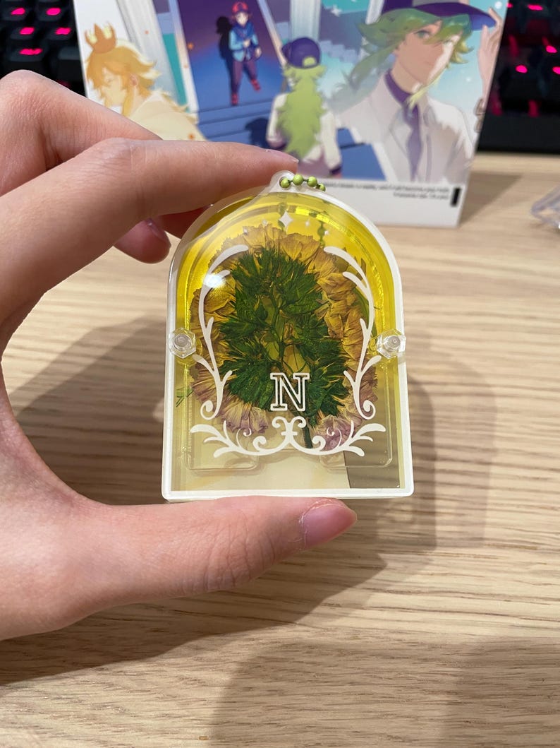 May include: A yellow and white rectangular keyring with a floral design and the letter "N". The keyring has a clear centre with dried flowers and decorative flourishes. The background shows an anime-style illustration.