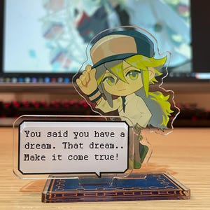 May include: A clear acrylic stand featuring a cartoon character with yellow hair and a hat. The character is holding up a hand. A speech bubble reads, "You said you have a dream. That dream.. Make it come true!" The base has a blue design.
