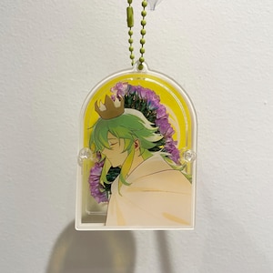May include: Acrylic keychain with an anime-style character with green hair and a gold crown. The character is set against a yellow backdrop with purple floral accents. The keychain hangs from a green beaded chain.