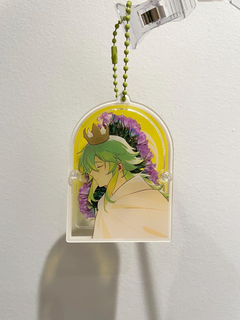 May include: Acrylic keychain with an anime-style character with green hair and a gold crown. The character is set against a yellow backdrop with purple floral accents. The keychain hangs from a green beaded chain.