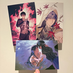 May include: Three rectangular art prints featuring anime-style illustrations. One shows a person in a suit surrounded by pink flames, another depicts a hooded person with purple hair and red leaves, and the third shows two figures embracing in water.