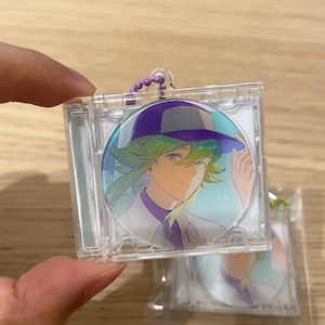 May include: A clear plastic keychain with a circular image of an anime character with green hair and a purple hat. The character is wearing a white shirt and purple collar. The keychain has a purple beaded chain.