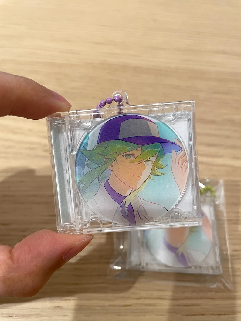 May include: A clear plastic keychain with a circular image of an anime character with green hair and a purple hat. The character is wearing a white shirt and purple collar. The keychain has a purple beaded chain.