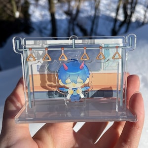 Puede incluir: Una pantalla acrílica transparente con un personaje de estilo anime de pelo azul con cuernos y alas, de pie en una escena de vagón de tren en miniatura. La pantalla tiene un marco transparente con una barra para colgar y asideros naranjas.