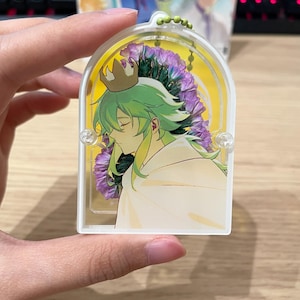 May include: A clear acrylic keyring featuring an anime-style character with green hair and a gold crown, set against a backdrop of purple flowers and a yellow background. The keyring has a small green beaded chain.