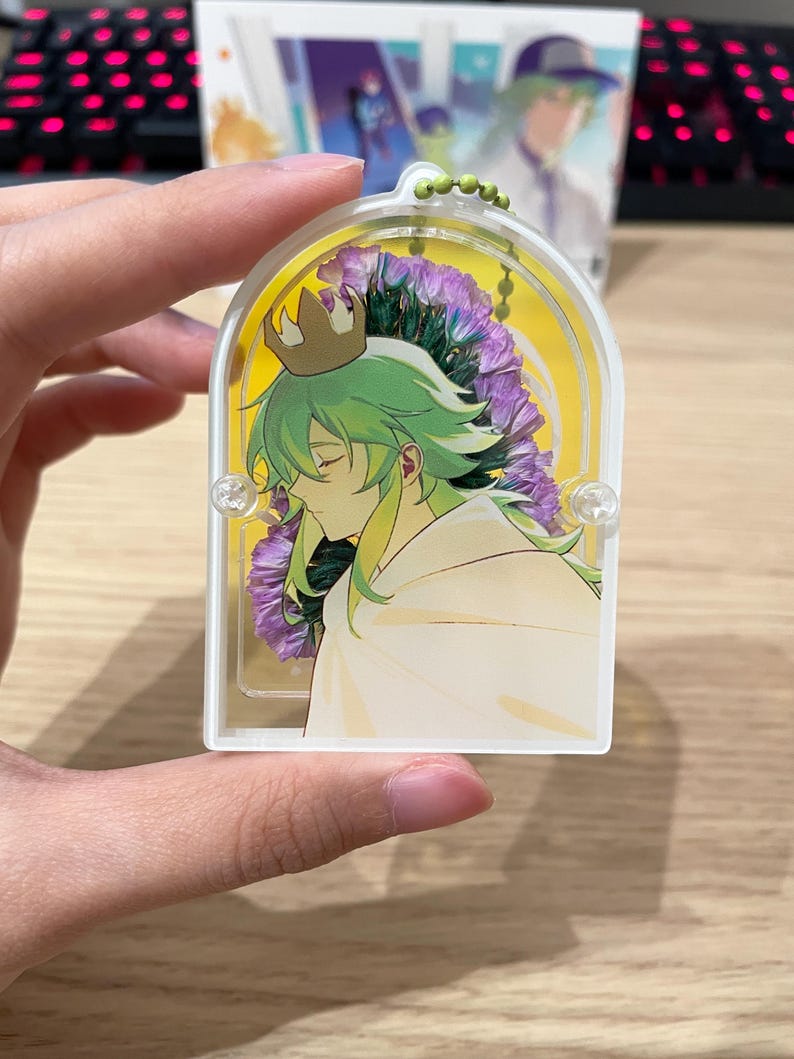 May include: A clear acrylic keyring featuring an anime-style character with green hair and a gold crown, set against a backdrop of purple flowers and a yellow background. The keyring has a small green beaded chain.