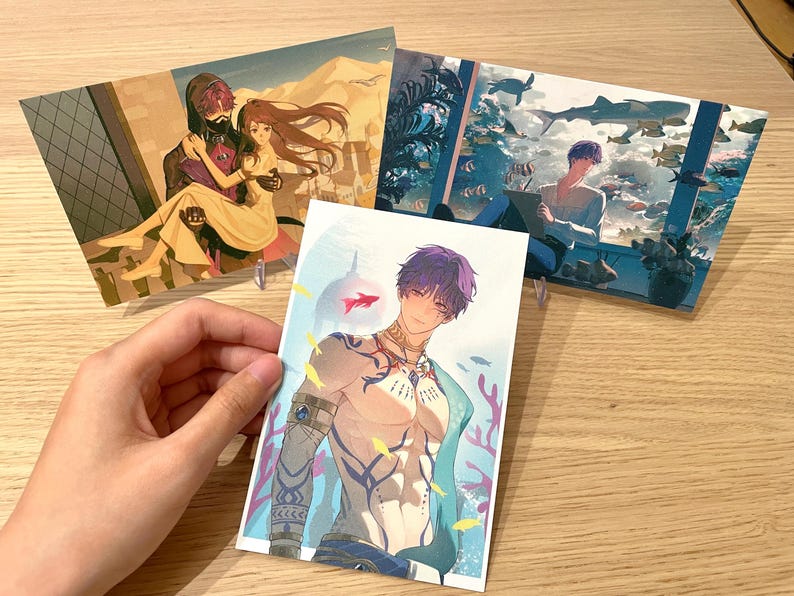 May include: Set of three art prints featuring anime-style illustrations. One print depicts a couple in a desert setting, another shows a person in an aquarium, and the third showcases a shirtless male figure with purple hair and aquatic elements.