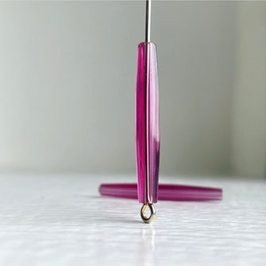 May include: A close-up of a pink glass bead with a gold metal loop. The bead is long and narrow, with a faceted surface.