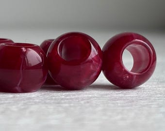 Pode incluir: Uma coleção de contas esféricas num tom bordeaux profundo e rico com um acabamento brilhante e marmoreado. Cada conta apresenta um orifício central para enfiar, exibindo uma superfície lisa e polida com variações subtis de tom.