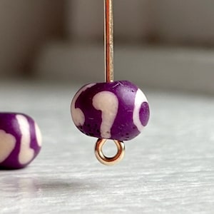 May include: Close-up of two purple and white beads. One bead is attached to a gold-colored wire, while the other bead rests separately. The beads have a rounded shape with a pattern of white swirls against a purple background. The beads are likely for jewelry making.