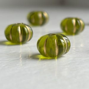 May include: A collection of small, round, translucent olive green glass beads. Each bead features a distinct ribbed or melon-like texture with vertical metallic gold-toned stripes running along the sides, creating a reflective and segmented appearance.