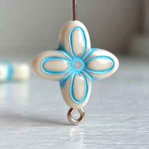 May include: A cream-colored, four-petal flower bead with a light blue outline and center detail. The bead is suspended by a thin wire, and a small silver loop is attached at the bottom. Another bead with a similar design is blurred in the background.