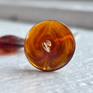 May include: Close-up of two decorative buttons. The primary button is a translucent amber color with a gold-colored shank. The second button is a smaller, dark red, teardrop shape. Both buttons are on a light-colored surface.