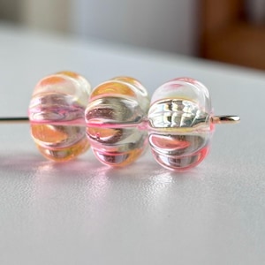 Puede incluir: Tres cuentas de vidrio transparente con un diseño acanalado, ensartadas en un alambre dorado. Las cuentas tienen una mezcla de tonos rosa, naranja y amarillo, creando un efecto colorido y translúcido. Las cuentas son probablemente para la fabricación de joyas.