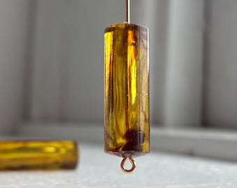 Op de afbeelding: Een cilindervormige, amberkleurige kraal met interne wervelpatronen, hangend aan een dunne metalen draad. De kraal heeft een klein metalen oogje aan de onderkant voor bevestiging. Een tweede identieke kraal ligt horizontaal op de achtergrond.