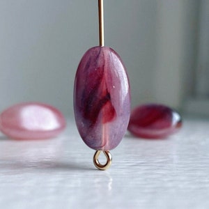 May include: A close-up of a purple and pink oval-shaped glass bead with a gold loop at the bottom. The bead has a swirl pattern and is translucent.