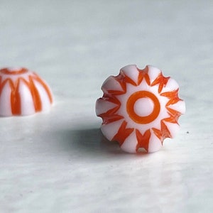 May include: Two white and orange glass buttons. One button has a striped pattern and the other has a sunburst design.