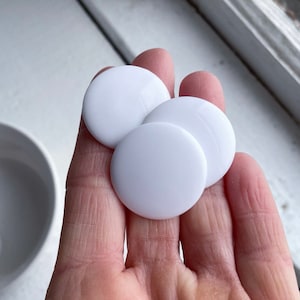May include: Three white round buttons, approximately 1 inch in diameter, are held in a hand. The buttons are smooth and have a glossy finish.