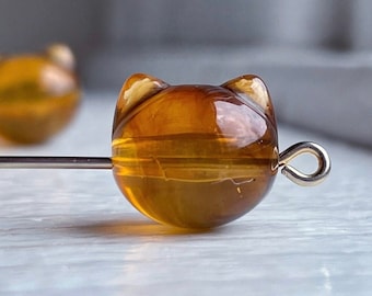 Pode incluir: Close-up de uma conta em forma de gato com um tom dourado. A conta está presa a um pino de metal prateado com um laço. Outra conta semelhante está ligeiramente desfocada no fundo.