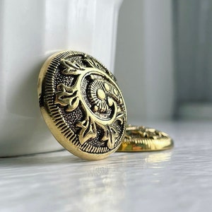 May include: Two gold-toned metal buttons with an ornate design. The buttons have a raised, detailed pattern of leaves and swirls.
