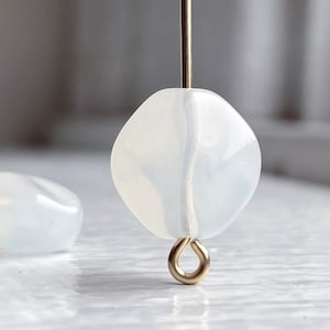 May include: Two white, translucent gemstone beads. One bead is suspended on a gold-colored wire, and the other rests on a white surface. The beads have a rounded, square shape. The background is blurred.