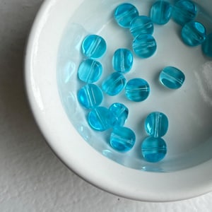 May include: A collection of translucent, round, light blue beads with a white stripe detail, resting in a white ceramic bowl. The beads are partially submerged in a light blue liquid, creating a reflective surface.