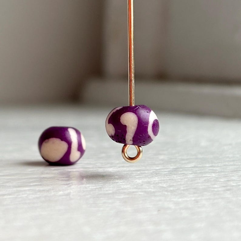 May include: Close-up of two purple and white beads. One bead is attached to a gold-colored wire, while the other bead rests separately. The beads have a rounded shape with a pattern of white swirls against a purple background. The beads are likely for jewelry making.