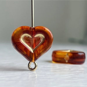 May include: A heart-shaped glass bead with a gold heart design inside. The bead is brown and amber in color and has a gold loop at the bottom.