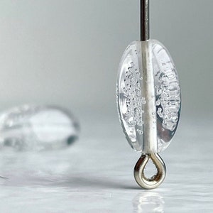 Puede incluir: Una cuenta de vidrio transparente con un bucle plateado y pequeñas burbujas de aire en todo. La cuenta está enhebrada en un alambre plateado.