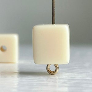 May include: A close-up of a white square bead with a gold loop on the bottom. The bead is on a white surface.