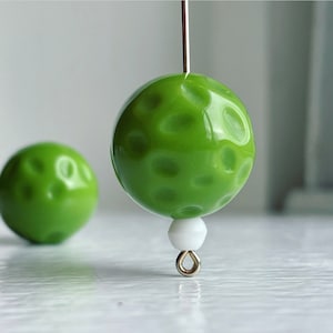 Può includere: Una perla sferica verde lime brillante con una superficie testurizzata e punteggiata. La perla è infilata su un sottile perno in metallo argentato, con una piccola perlina distanziatrice bianca e un occhiello in metallo argentato alla base. Una seconda perla identica è fuori fuoco.