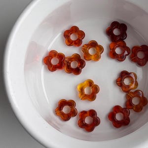 May include: A collection of translucent flower-shaped beads in shades of amber and brown, resting in a white bowl. The beads have a five-petal design with a central hole, ideal for jewelry making or craft projects.