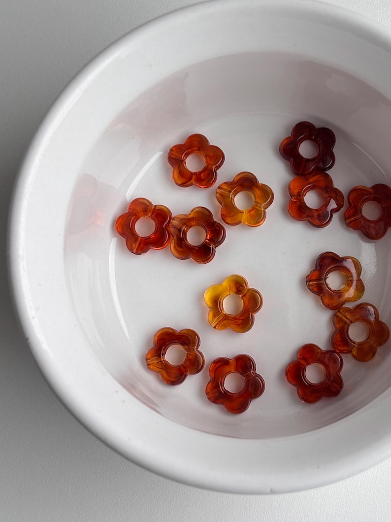 May include: A collection of translucent flower-shaped beads in shades of amber and brown, resting in a white bowl. The beads have a five-petal design with a central hole, ideal for jewelry making or craft projects.