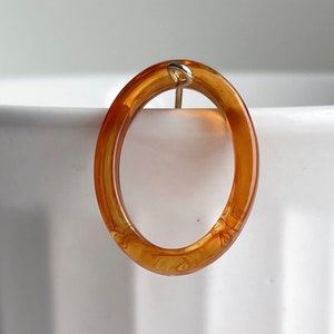 May include: A single, oval-shaped earring with a warm amber hue. The earring is suspended from a small, gold-colored loop. The transparent material reveals subtle patterns within the amber, creating a unique visual texture. The earring is set against a soft, white background.