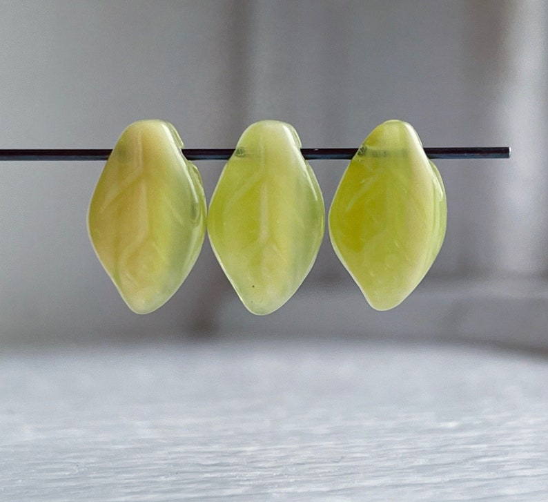 Op de afbeelding: Drie lichtgroene glazen kralen in de vorm van een blad, geregen aan een zwarte draad.
