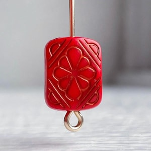 May include: A red, rectangular bead with a floral design carved into its surface. The bead is suspended by a gold-colored wire and a gold-colored loop. The bead's design features a central flower and geometric patterns.