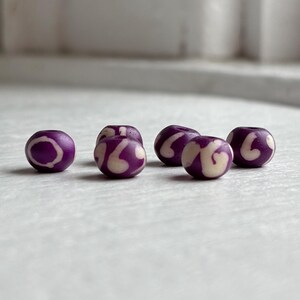 May include: Five round purple beads with a cream-colored swirl design. The beads are evenly spaced and appear to be made of a smooth material. The background is a light, neutral color.