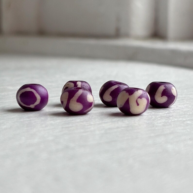 May include: Five round purple beads with a cream-colored swirl design. The beads are evenly spaced and appear to be made of a smooth material. The background is a light, neutral color.