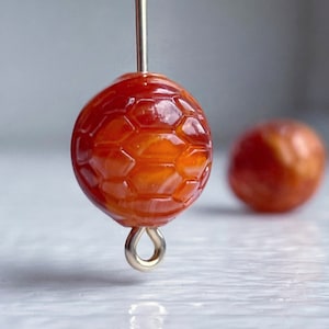 May include: A close-up of a round, orange, and yellow bead with a honeycomb pattern. The bead is attached to a gold loop.