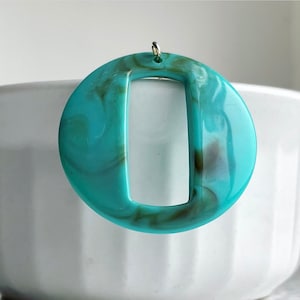 May include: A turquoise and brown marbled resin earring with a silver-colored metal loop at the top. The earring has a circular shape with a rectangular cutout in the center. The earring is set against a white background.