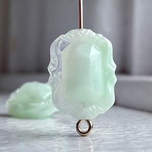 May include: A pale green glass bead with a decorative, ornate design. The bead has a gold-toned metal loop attached to the bottom.