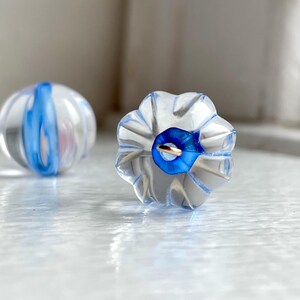 以下が含まれることがあります： 青と白の渦巻き模様が入った透明なガラスビーズが2つ。1つは丸型、もう1つは花の形をしています。