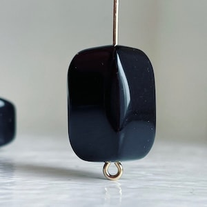 Puede incluir: Cuente negra de forma cuadrada con un aro de salto dorado. La cuenta tiene un acabado brillante y pulido.